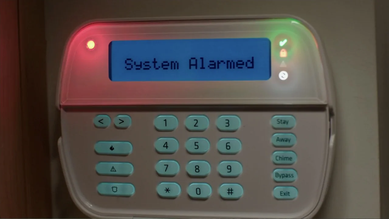The Intern (2015) movie still: closeup — A white home security alarm panel is mounted on a plain wall. The blue digital display shows the text…