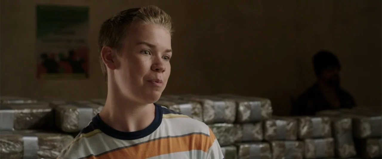 We're the Millers (2013) movie still: medium — A dimly lit warehouse interior features stacks of plastic-wrapped parcels in the background. A young…