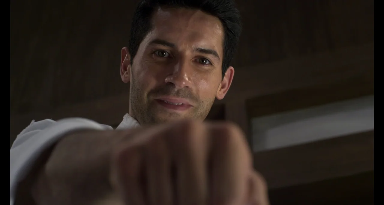 Ninja: Shadow of a Tear (2013) movie still: closeup — A dimly lit room with dark wooden paneling in the background. A man reaches his arm forward…