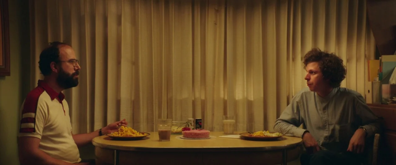 Lemon (2017) movie still: medium — A small dining room features a round table centered against a wall of pleated curtains. Two men sit at the table…