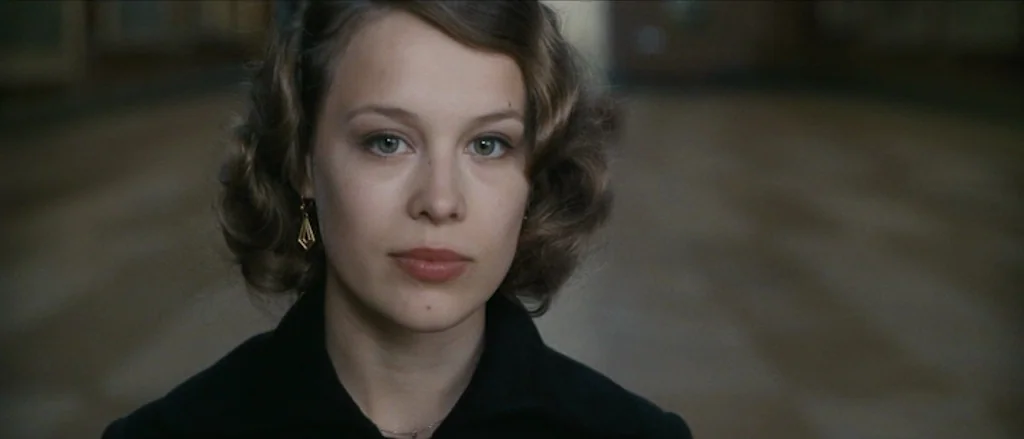 Frantz (2016) movie still: closeup — A woman stands in a blurred, indistinct hallway with wooden floors. She faces the camera directly with a…