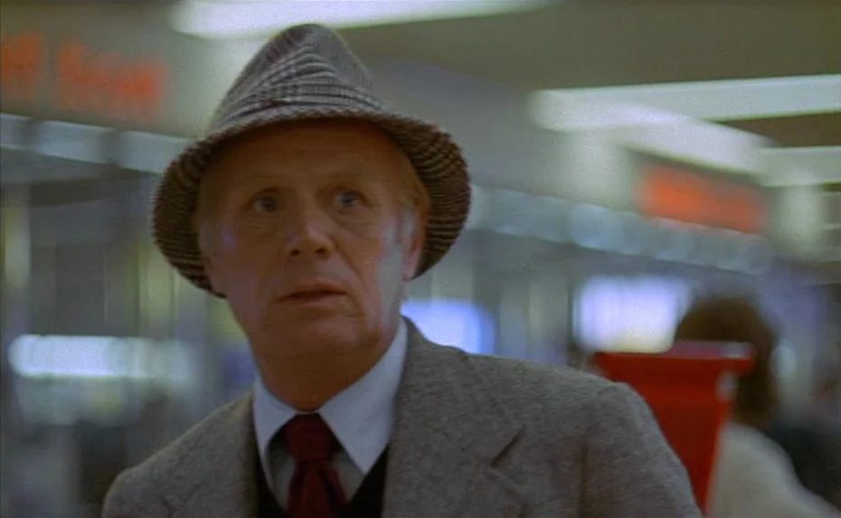 To The Devil a Daughter (1976) movie still: medium — An older man stands inside a brightly lit retail space with blurred shelves behind him. He…