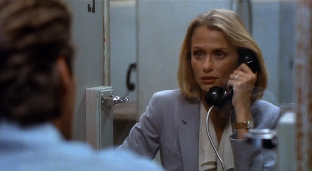 American Gigolo (1980) movie still: medium — A spartan room features gray metal partitions with telephone units for communication. A woman in a…