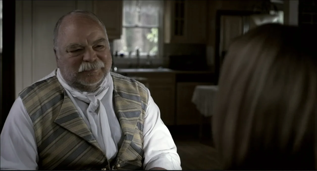 The Secret Village (2013) movie still: medium — A residential kitchen with wooden cabinets and a blurred window in the background. An older man in a…