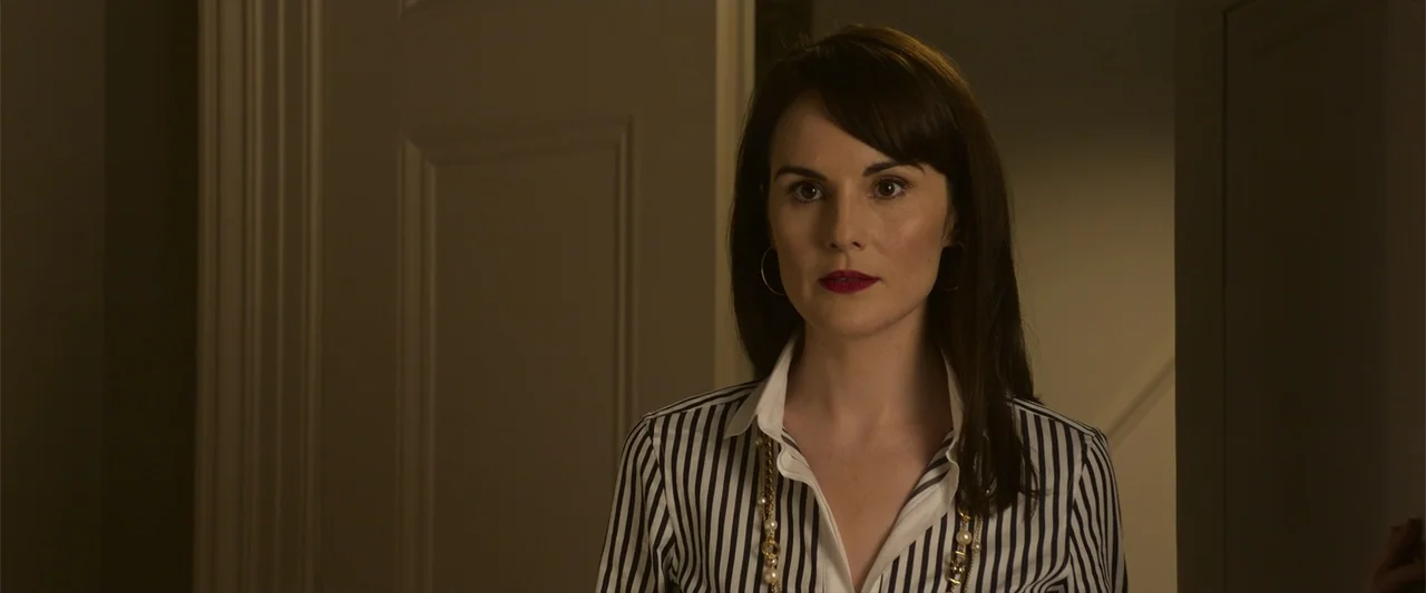 The Gentlemen (2020) movie still: medium — A woman stands in a domestic hallway with beige walls and a white door frame. She is looking directly…