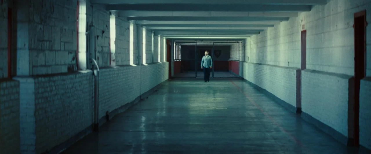 Out of the Furnace (2013) movie still: wide — A long, industrial prison hallway features painted white brick walls and a concrete floor. A single…