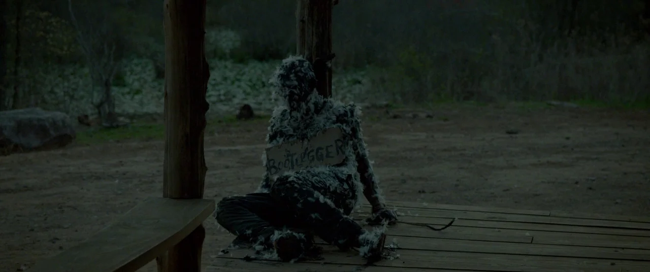 Lawless (2012) movie still: medium — A dark wooden porch in a rural setting during late evening. A person covered in tar and feathers sits against a…
