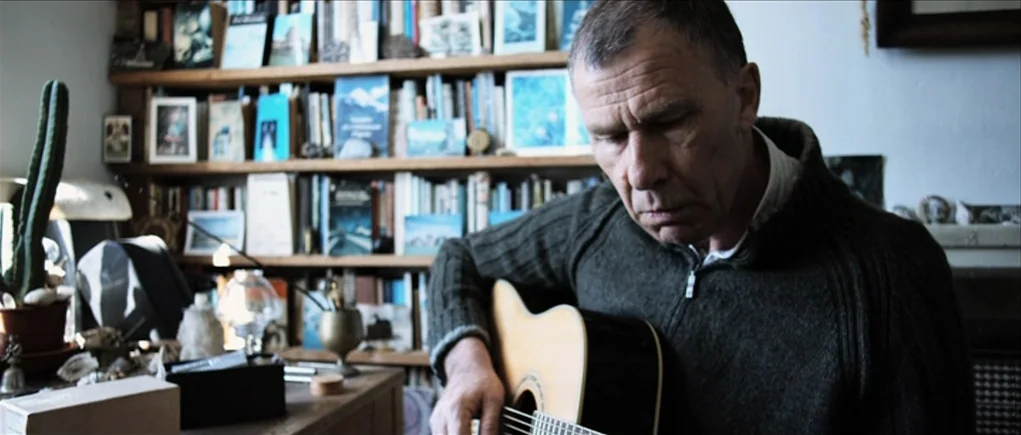 Down Terrace (2009) movie still: medium — A man sits in front of a floor-to-ceiling bookshelf filled with books and small objects. He plays a wooden…