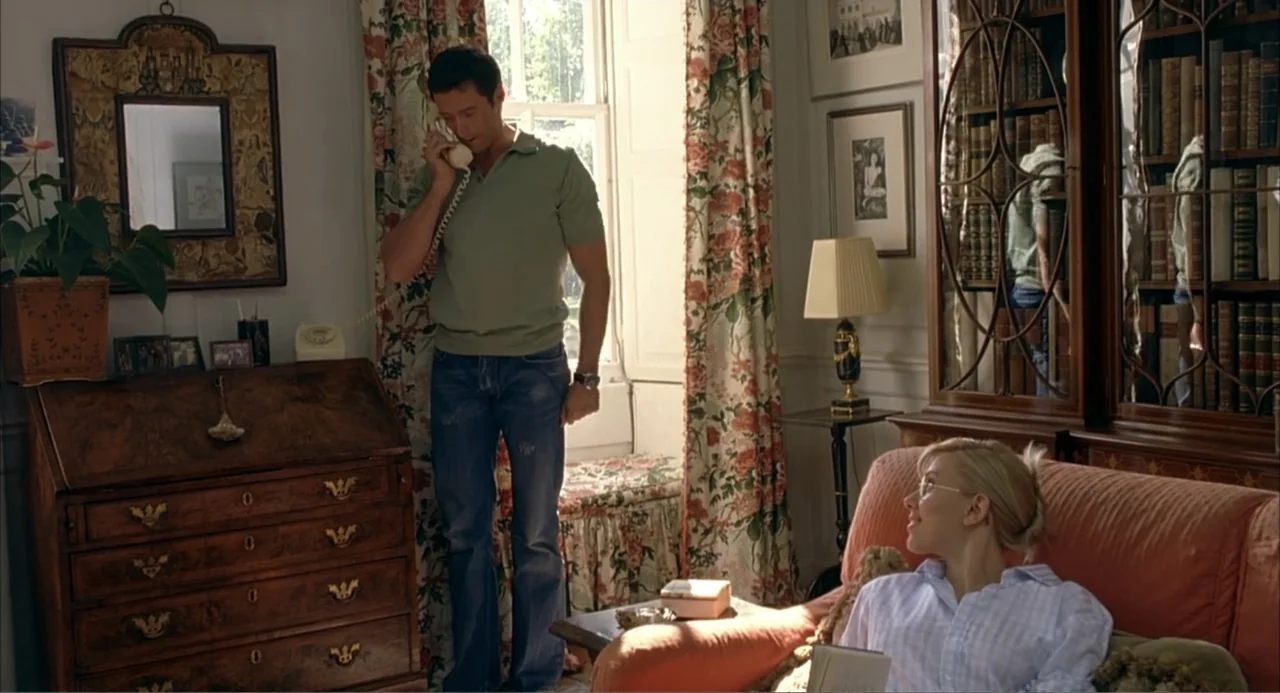 Scoop (2024) movie still: medium — A furnished interior features a wooden writing desk, a floral couch, and a bookshelf. A man talks on a vintage…