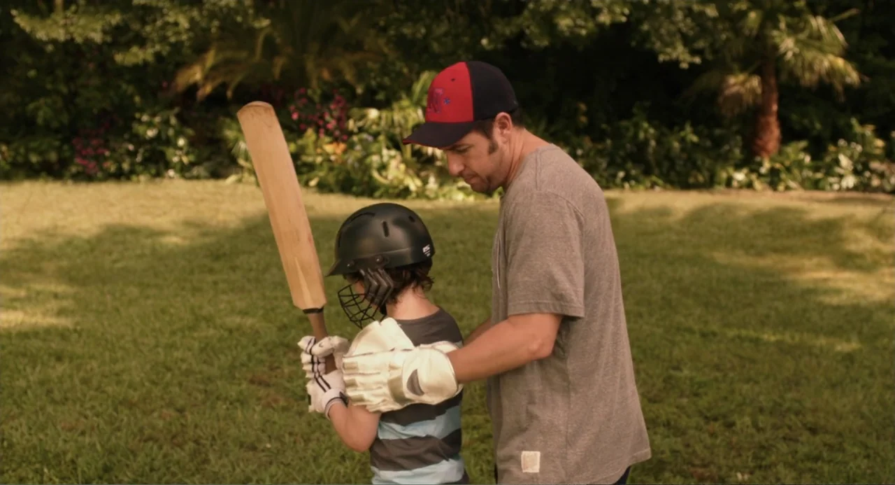 Blended (2014) movie still: medium — A grassy lawn with mature trees and foliage in the background during the day. An adult male stands behind a…