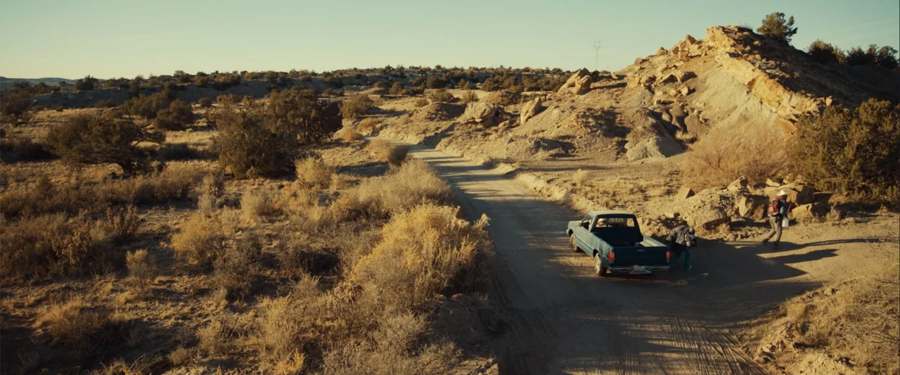 Frontera (2014) movie still: wide — A dry desert landscape with scrub brush and rocky hills surrounds a narrow road. Two people stand beside a teal…