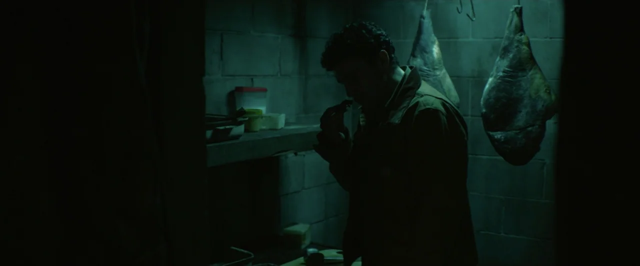 Wolf Man (2025) movie still: medium — A dimly lit room features cinder block walls and large cuts of meat hanging. A man in a work jacket stands…