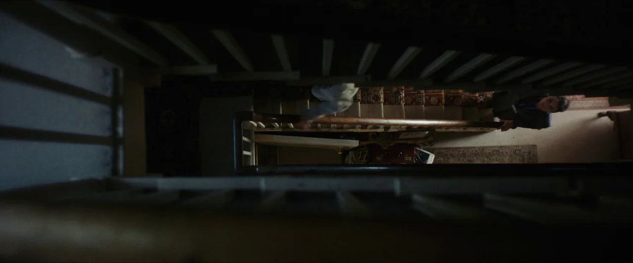 Fréwaka (2024) movie still: wide — A top-down view looks down a wooden staircase with a central bannister rail. The stairs are covered with dark…