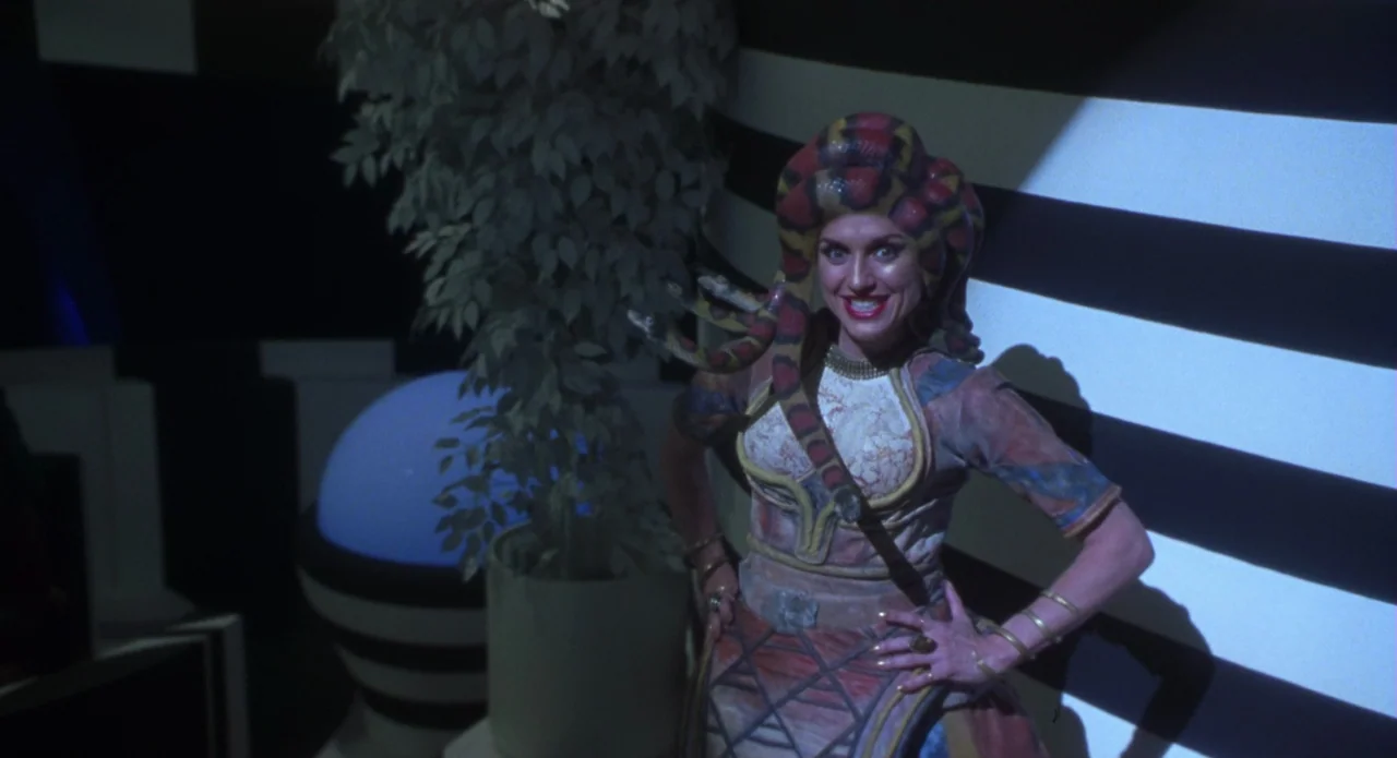 Monkeybone (2001) movie still: medium — A woman stands in a surreal, minimalist interior with high-contrast black and white striped walls. She wears…