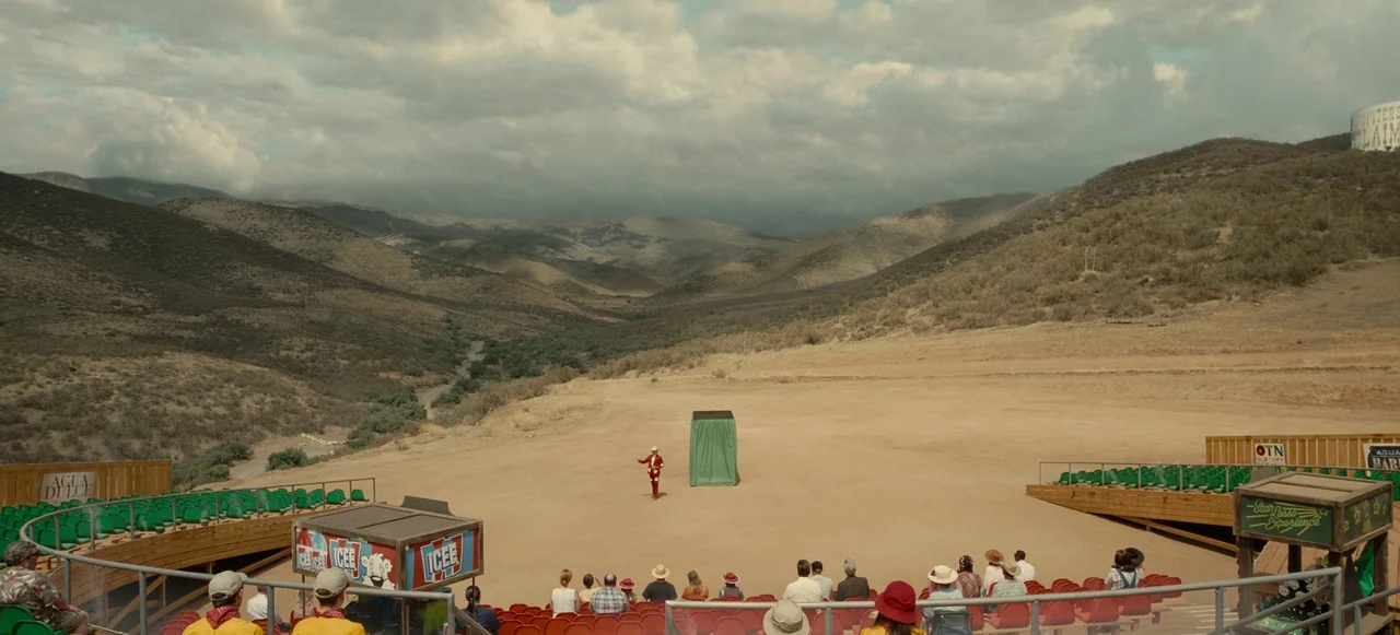 Nope (2022) movie still: wide — A dry, hilly valley landscape serves as the backdrop for a small outdoor stage. A man in a red suit stands alone on…