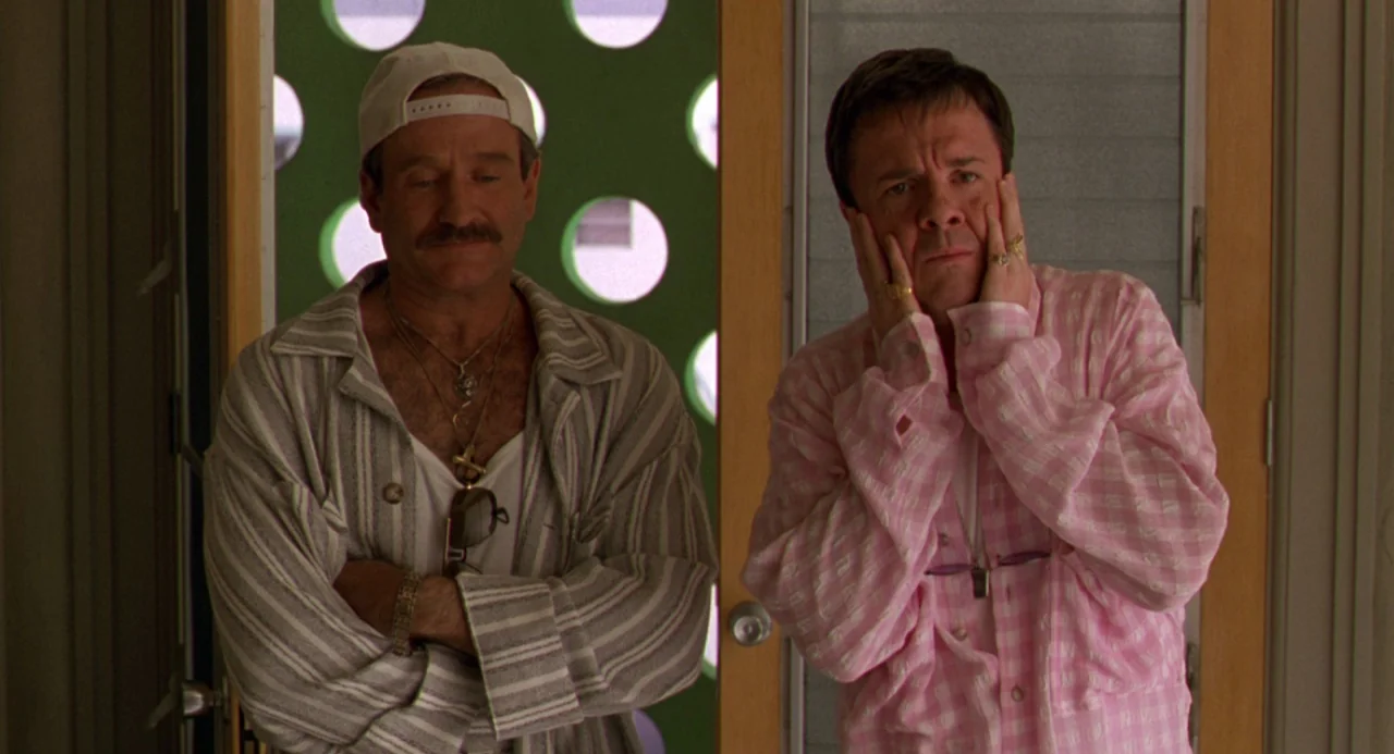 The Birdcage (1996) movie still: medium — Two men stand in front of a green door featuring circular cutouts. They are positioned side-by-side facing…