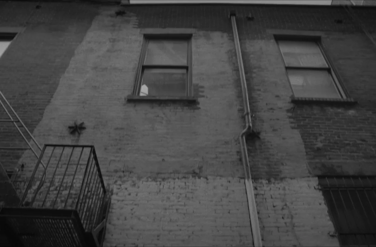 The Kitchen (2012) movie still: establishing — The exterior brick wall of a multistory building features two windows and a metal fire escape. A…