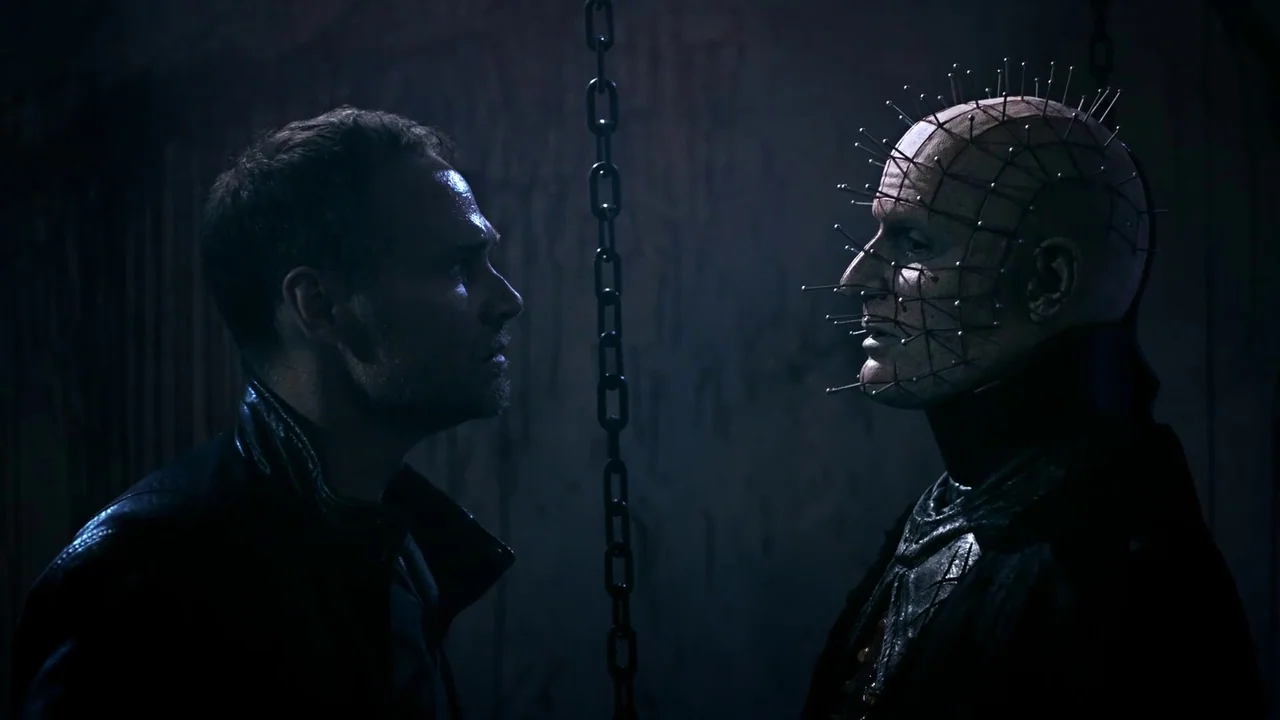 Hellraiser: Judgement (2018) movie still: medium — A dark, cramped interior with a heavy metal chain hanging vertically between two men. One man…