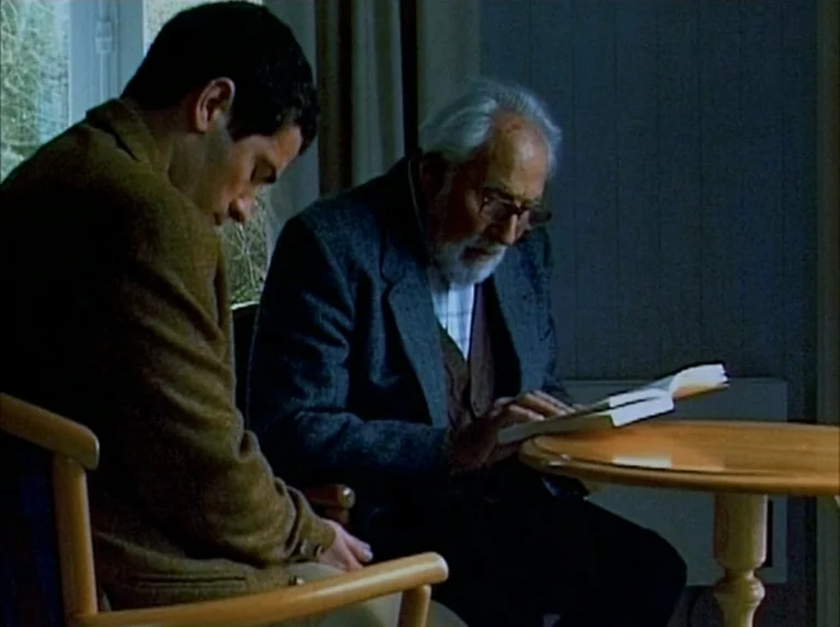 Eloge de L’amour (2001) movie still: medium — A quiet residential room features a circular wooden table in front of two seated men. The older man…