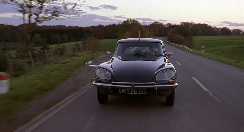 CQ (2001) movie still: medium — A rural paved road winds through flat fields under a dim evening sky. A black Citroën DS drives toward the camera on…