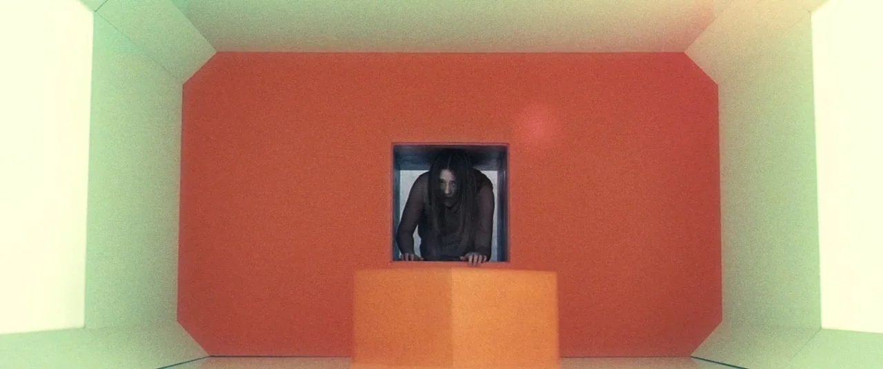 Beyond The Black Rainbow (2010) movie still: wide — A symmetrical room has vibrant orange central walls and pale green side panels. A woman looks…