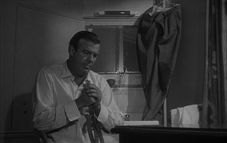 Le Signe du Lion (1962) movie still: medium — A dark bathroom contains a sink and a mirror hanging on the wall. A man sits in a chair while looking…