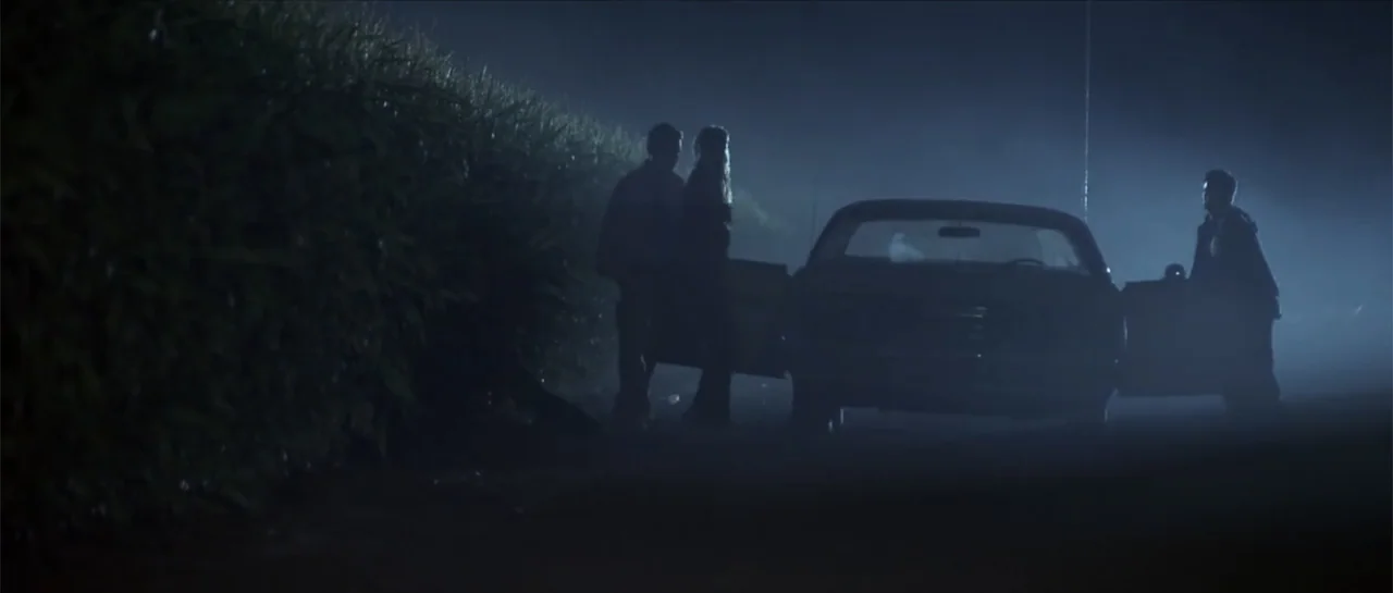Joy Ride (2001) movie still: wide — A rural road at night borders a dense field of tall crops under fog. Three figures stand beside a sedan with its…