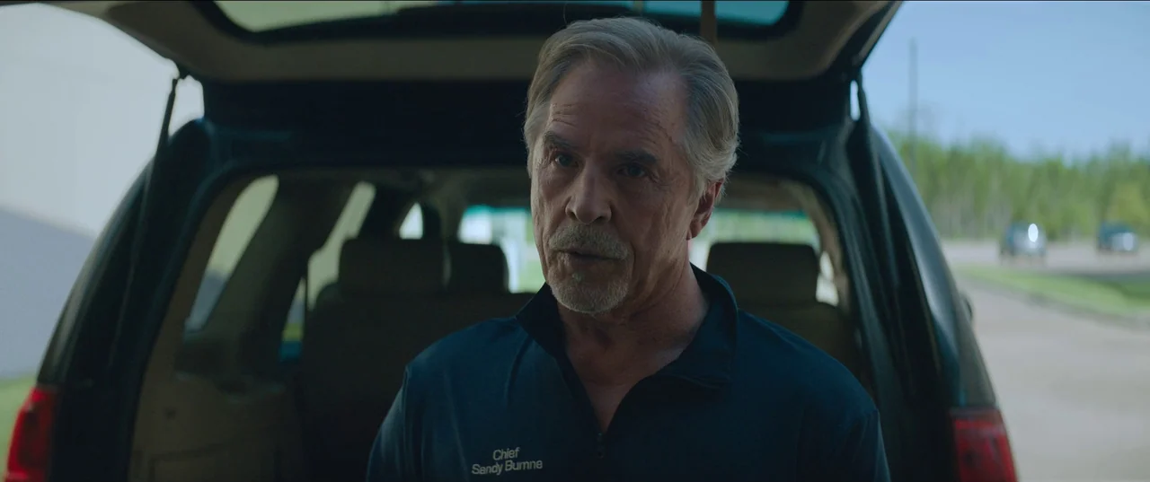 Rebel Ridge (2024) movie still: medium — A man stands before the open hatch of an SUV parked in an outdoor lot. He is wearing a dark uniform shirt…