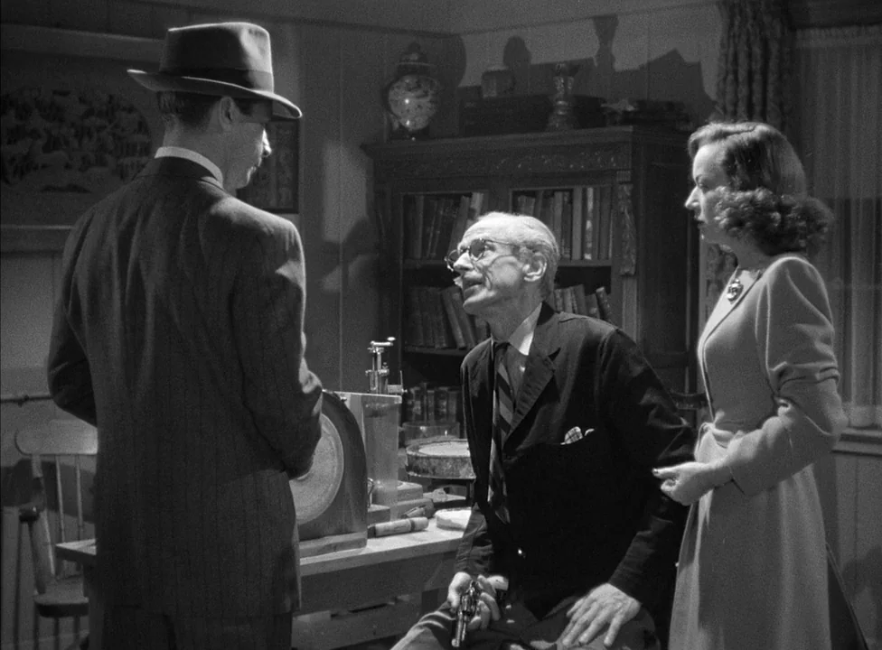 Murder, My Sweet (1944) movie still: medium — A dark study contains bookshelves, a wooden table, and a desk. An older man seated at a table holds a…