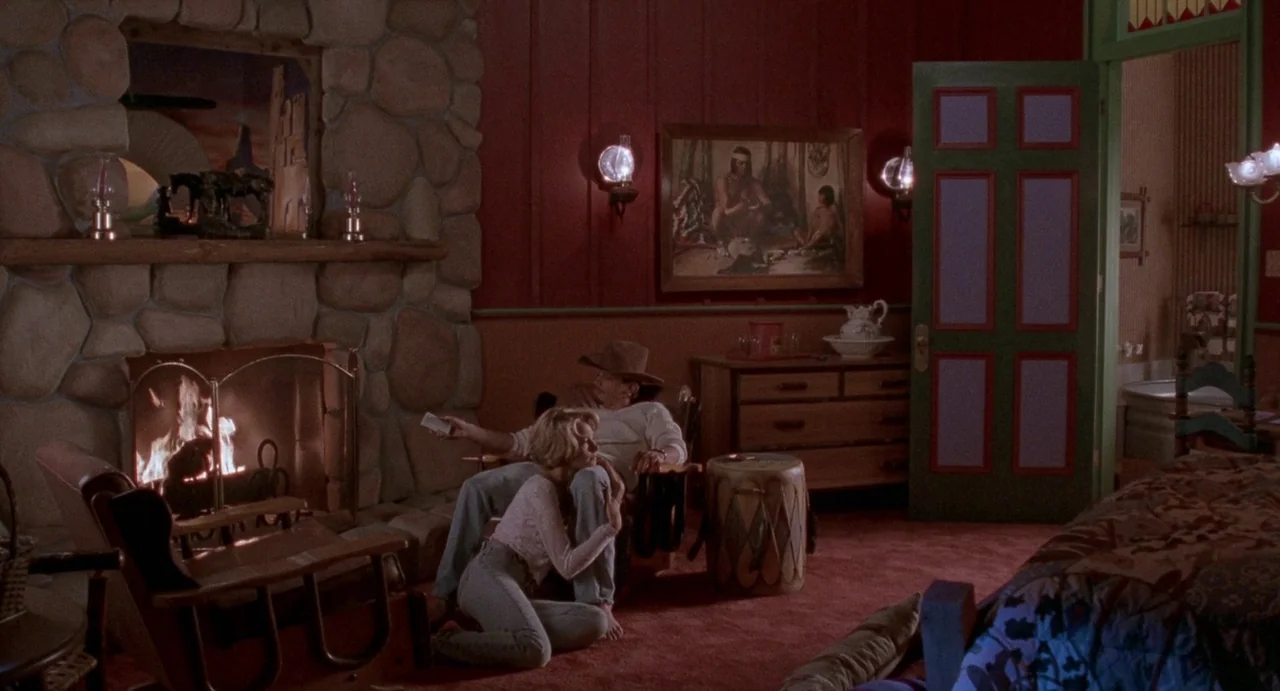 Touch (1997) movie still: wide — A rustic interior with red walls and a large stone fireplace mantle. A man sits in a wooden rocking chair while a…