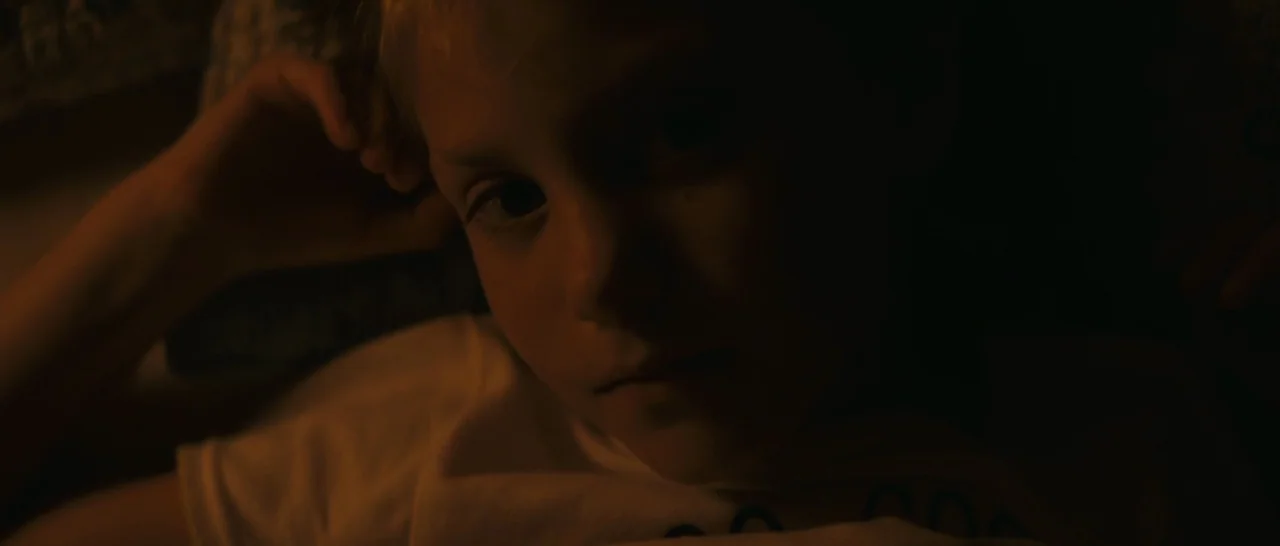 Arrival (2016) movie still: closeup — A dark, dimly lit indoor space with the subject resting on a soft surface. A young child lies with their head…