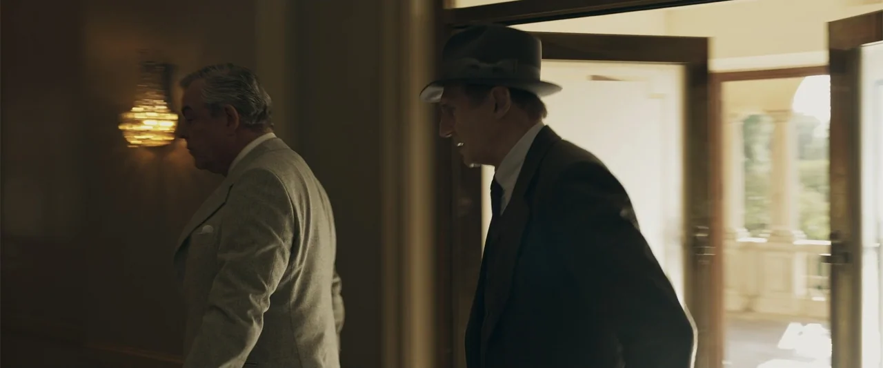 Marlowe (2023) movie still: medium — An interior hallway features a doorway leading to a sunlit exterior area. Two men in formal suits walk in…