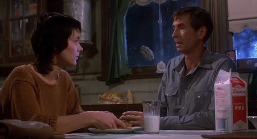 Psycho 2 (1983) movie still: medium — A dimly lit kitchen features a table with a carton of milk and glass. Two people sit facing each other at the…