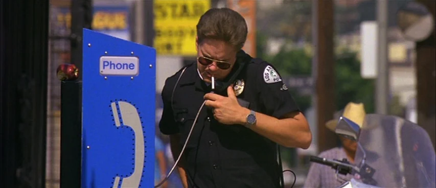 Short Cuts (1993) movie still: medium — A Los Angeles police officer stands on a sunny sidewalk next to a public phone. He holds a radio handset to…