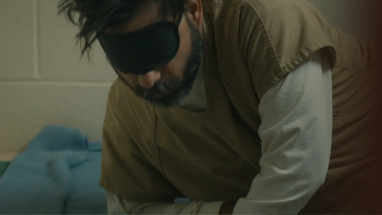 Camp X-Ray (2014) movie still: medium — A stark room with off-white cinder block walls and a blue blanket visible. A man wearing a blindfold and tan…