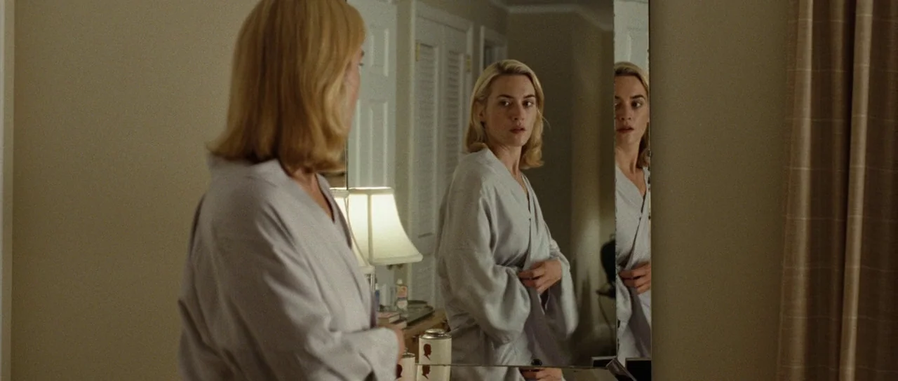 Revolutionary Road (2008) movie still: medium — A woman stands before a large wall mirror in a room with beige walls. She is adjusting her…