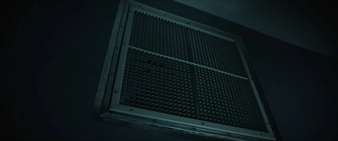 Vicious Fun (2021) movie still: closeup — A dark interior wall features a square white metal air vent cover. The vent slats are closed and recessed…