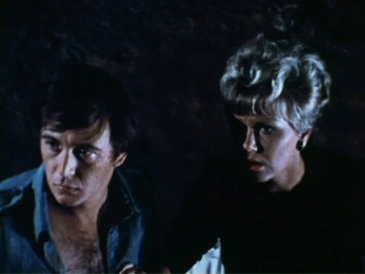 It's Alive (1974) movie still: medium — A dark indoor space with minimal background detail visible behind two people. A man and a woman look…