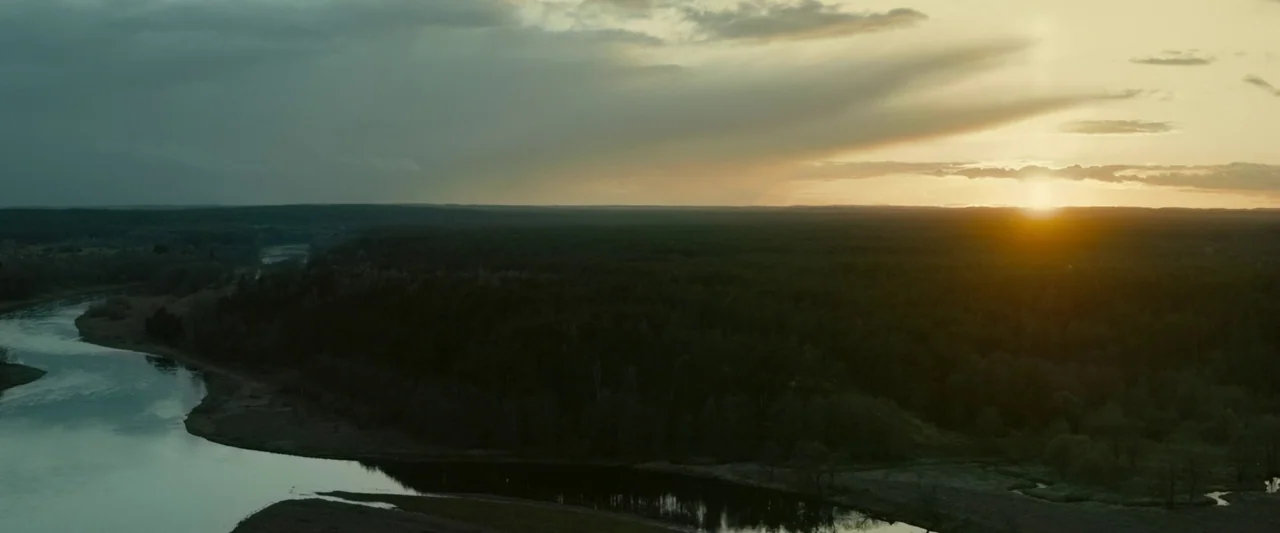 Vesper (2022) movie still: establishing — A vast landscape shows a winding river carving through a dense forest at dusk. The sun hangs low on the…