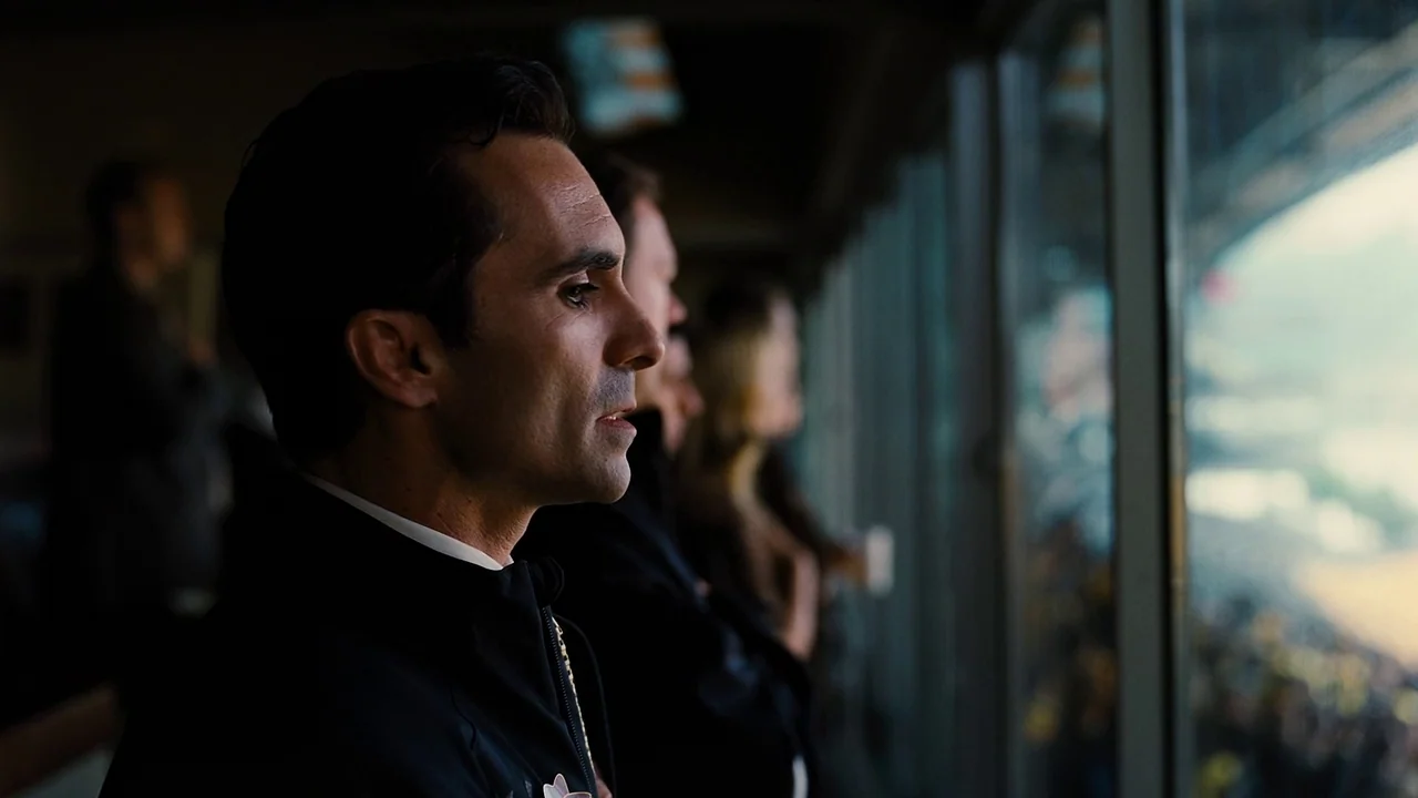The Dark Knight Rises (2012) movie still: medium — A luxury interior glass-walled viewing suite looks out over an active outdoor arena space. A man…