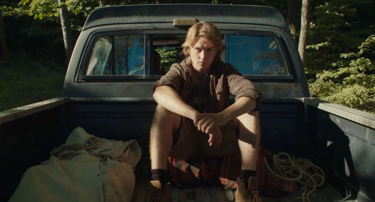 Cymbeline (2014) movie still: medium — A young man sits in the bed of an old, dark-colored pickup truck. The truck is positioned outdoors in a…