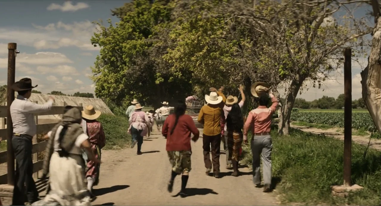 A Million Miles Away (2023) movie still: wide — A group of people walks away along a sunlit dirt road lined with trees. They wear casual clothing…