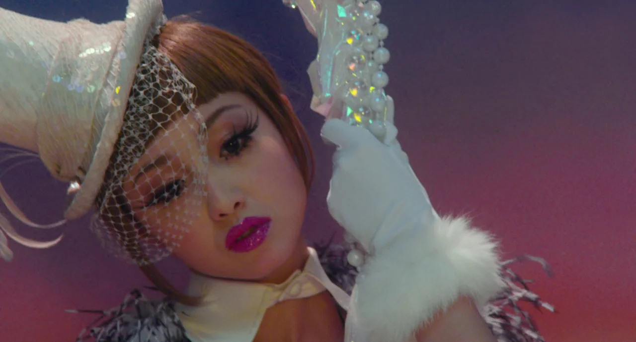 Helter Skelter (2012) movie still: closeup — A studio stage with a gradient background features a woman in formal costume. The woman holds an ornate…