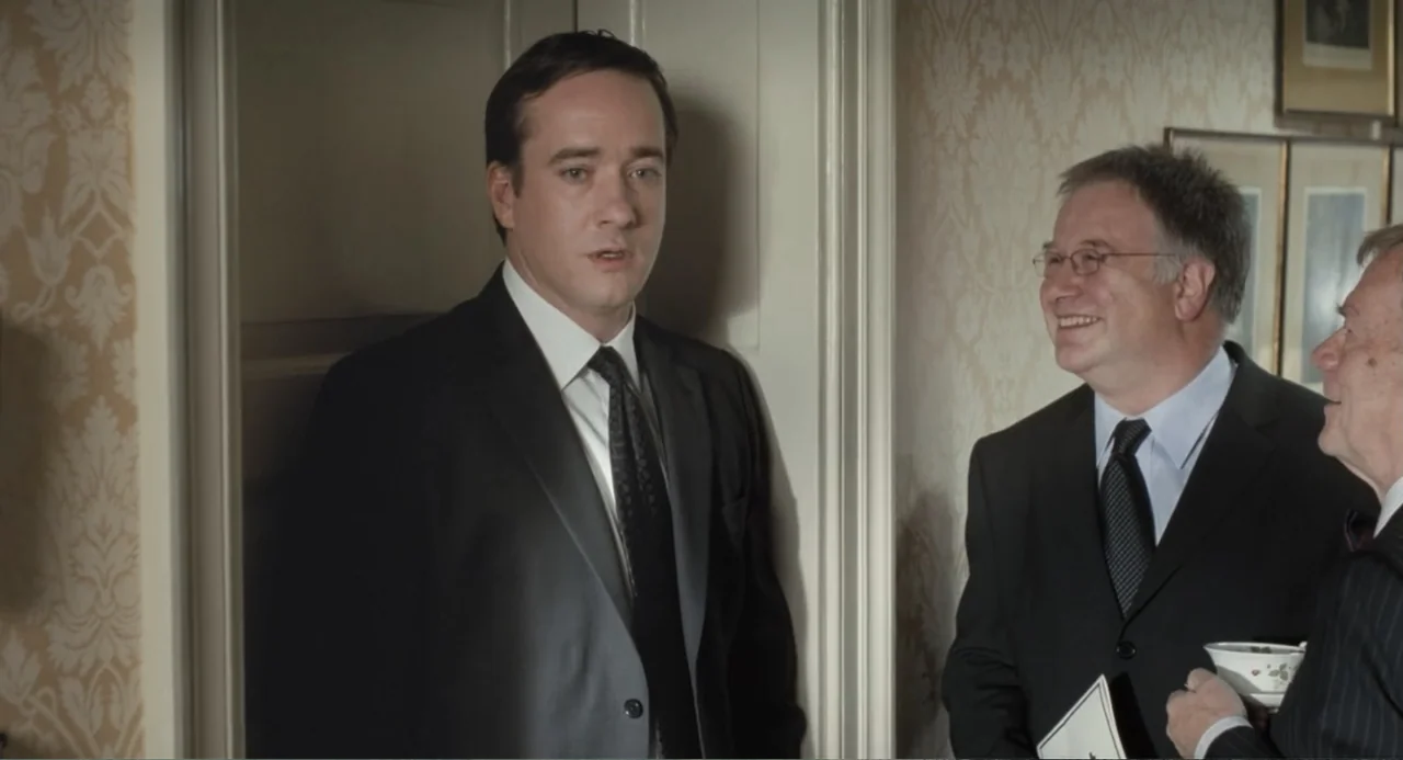 Death at a Funeral (2010) movie still: medium — A formal interior with patterned beige wallpaper and framed pictures on the wall. Three men in…