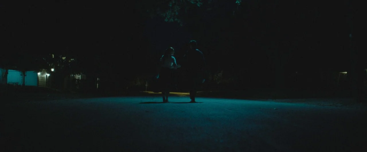 The Vast of Night (2019) movie still: wide — A dark residential street is illuminated by a single overhead light source at night. A young man and…