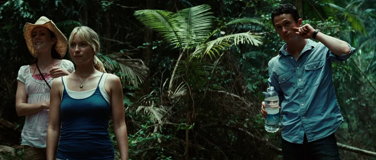 The Ruins (2008) movie still: medium — Three people stand within a thick, overgrown jungle environment surrounded by dense palm fronds. The group…