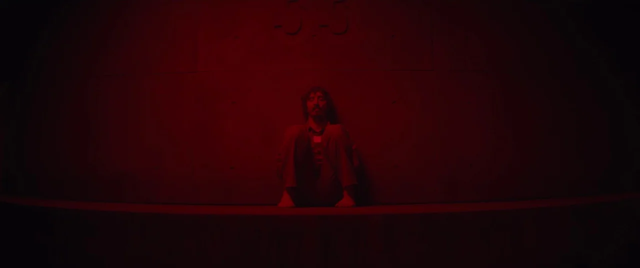 The Platform (2019) movie still: wide — A dark concrete room with a wall marked by the number 33 in relief. A man sits alone on the floor against…