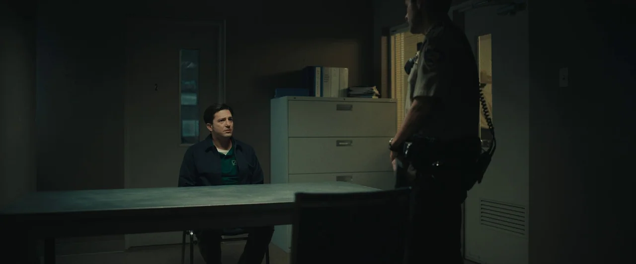 LaRoy, Texas (2023) movie still: wide — A dimly lit room contains a metal table and a filing cabinet against a wall. A man sits at the table as a…