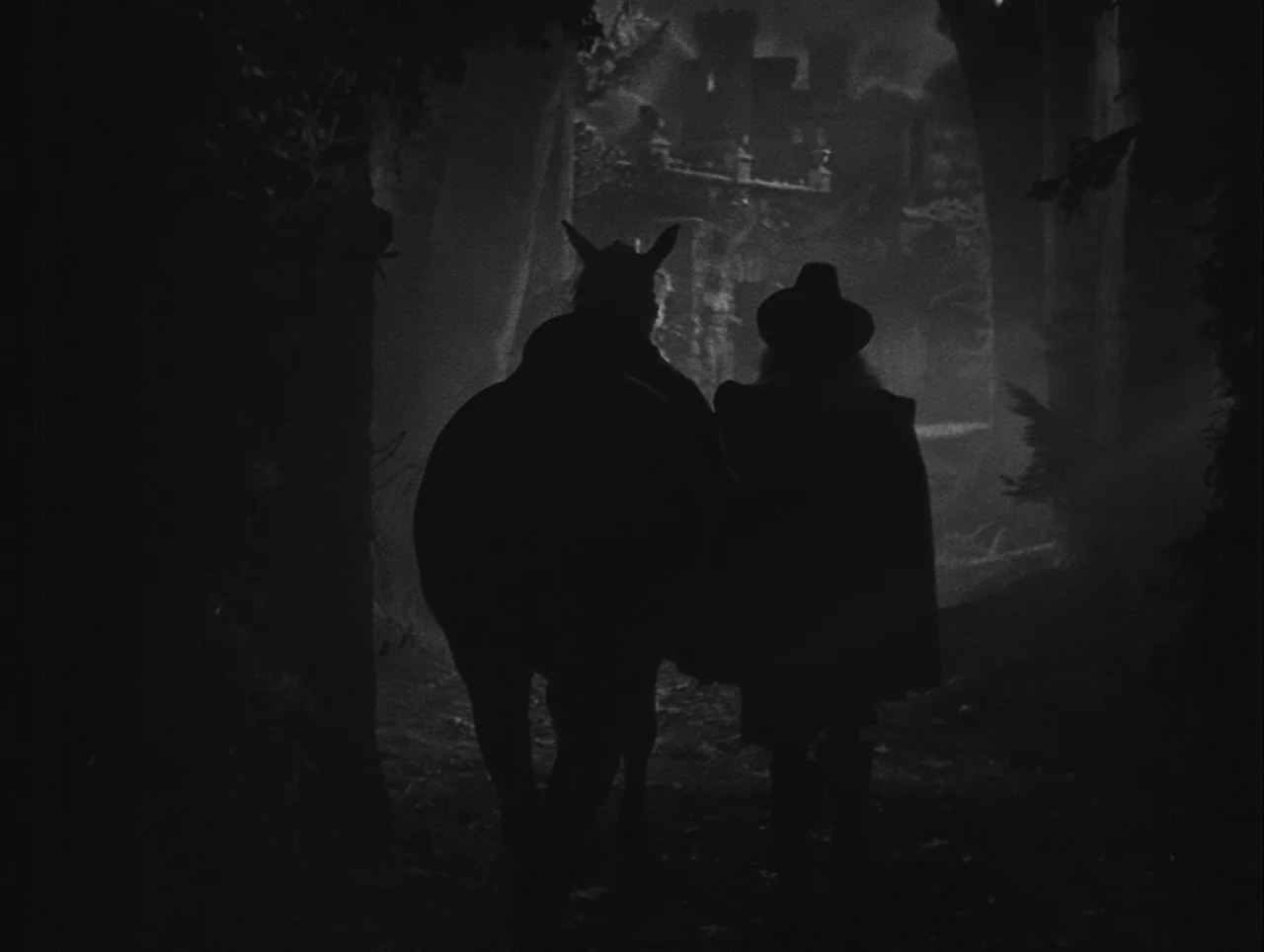 La Belle et la Bête (1946) movie still: fullbody — A dark, misty path is lined by tall trees with a distant castle visible ahead. A cloaked man…