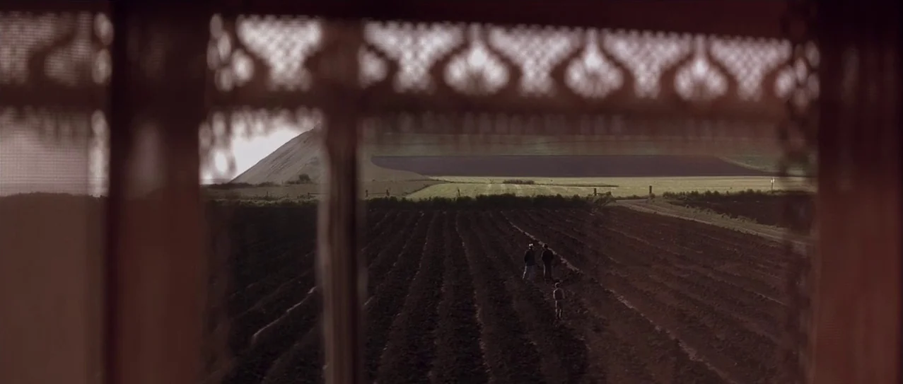 Snow Falling on Cedars (1999) movie still: wide — A vast, plowed agricultural field stretches toward a large hill under an open sky. Three figures…